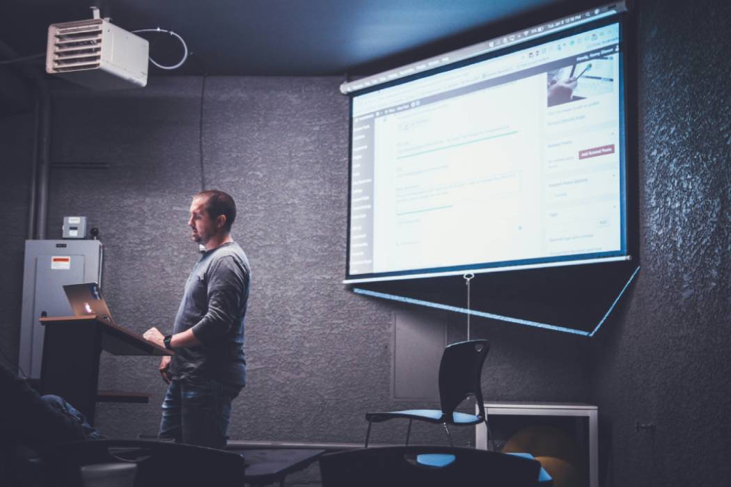 A person stading at a podium with a projector screen behind him.